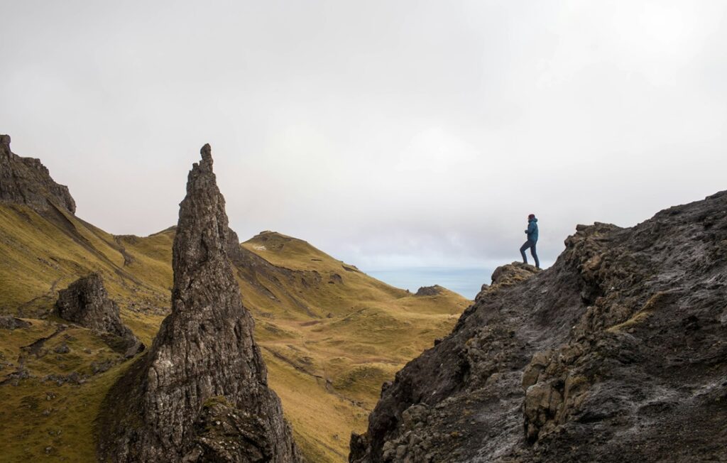 man stands at precipice of cliff face; gray skies above him